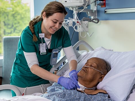 A female nurse holds hands with a male patient while smiling at him laying in his hospital bed.
