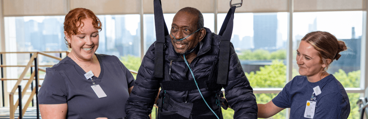 A patient utilizes equipment to support his weight and stand with the help of his therapists.